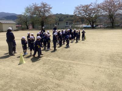 校庭のスタートラインに並ぶ1年生です。左手前から右奥に向かって先頭の児童が横に並んでおり、残りの児童が先頭の児童の後ろに並んでいます。担任と支援員が児童が並ぶ位置に個別に誘導しています。