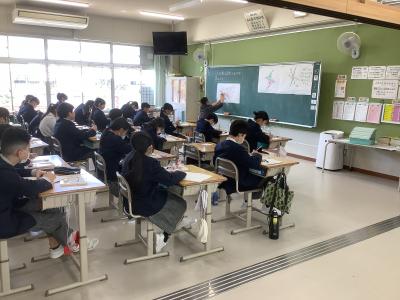 点対称な図形の作図を教える教師とそれをみる児童です。教室の右後方から対角線に写真を撮っています。黒板上に貼り付けた模造紙上に先生が指示を出しながら定規で線を引いています。児童はその指示を聞きながら、ノートに作図をしています。