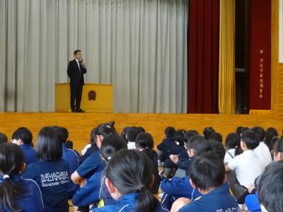 体育館の壇上でしゃべる校長の写真です。式台上のマイクスタンドからマイクを取り、手持ちで式台の前に立っています。画面の手前では児童が背中を向けて座っており、校長の方を向いています。