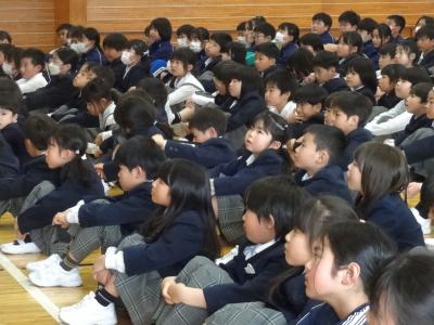 校長の話を聞く児童たちです。左側から取った写真で、半数以上の児童が校長の方へ顔を向けて座っていますが、一部の児童が顔を下に向けたり、上方を見上げたりしています。