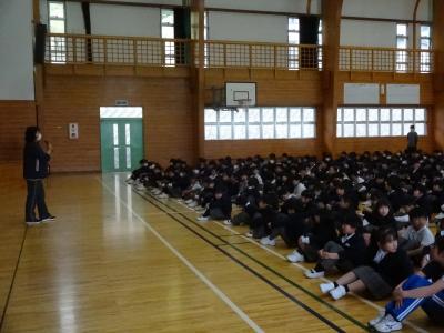 生徒指導の先生が児童の前で話している様子です。画面左で先生が話をして、右側の児童たちが座って話を聞いています。